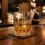 Gelas kristal berisi whisky warna amber keemasan dengan es bulat di atas meja bar kayu elegan